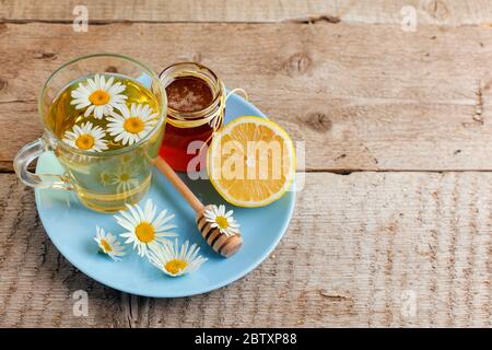 Tè sano camomilla versato in tazza di vetro. Teiera, piccolo vaso di miele, mazzo di erica e vaso di vetro di erbe medicinali a margherita. Foto Stock