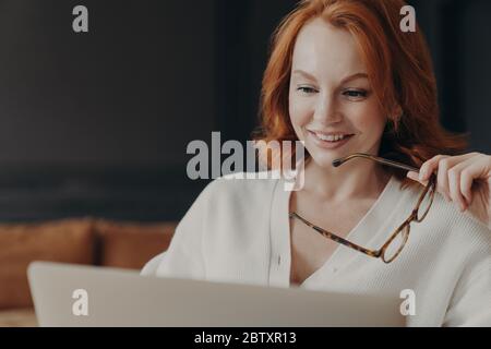 Bella donna con capelli rossi concentrata nel display del laptop, sorride positivamente, tiene occhiali ottici, posa in interni, utilizza le moderne tecnologie per il online Foto Stock