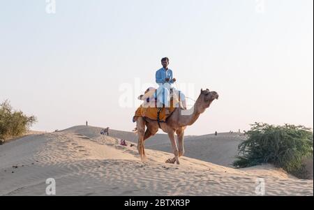 Un cammelliere (autista del cammello) che porta il cammello decorato mentre parla sui loro cellulari sulle dune di sabbia di sam del deserto di Thar con i turisti durante il tramonto Foto Stock