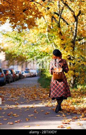 Elegante rosso femmina in plaid cappotto, berta nera e zaino camminare in città strada in giornata di sole, vista posteriore. Umore autunnale, concetto di fogliame. Rilassante Foto Stock