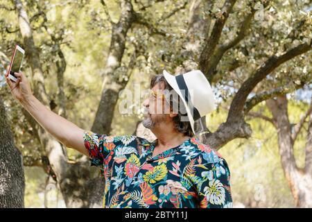 Profilo di un uomo con barba e cappello con una camicia con stampa floreale che fa un selfie con un cellulare in un giardino Foto Stock