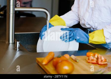 Dettaglio delle mani di una persona con guanti che lavano un piatto con acqua in un lavello da cucina con alcuni pomodori tagliati in un pannello di legno Foto Stock