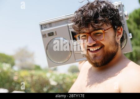 Giovane uomo grasso con un orecchio piercato e occhiali che indossa una cassetta radio sulla spalla mentre sorride all'aperto in una giornata di sole Foto Stock