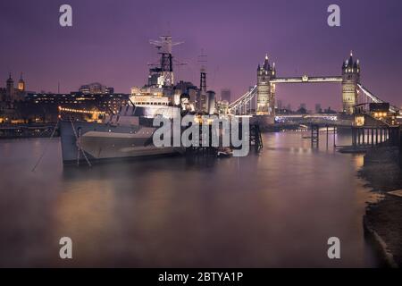 HMS Belfast con Tower Bridge in background, girato la mattina presto con Thames Mist e luci della città, Londra, Inghilterra, Regno Unito, Europa Foto Stock