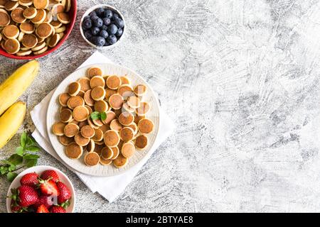 Minuscoli pancake per colazione. Frittelle di cereali con mirtilli, banane, fragole su sfondo grigio. Cibo alla moda. Spazio di copia per testo o disegno Foto Stock