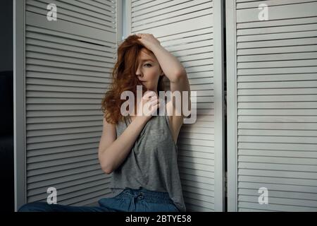 Ragazza rossa con i denti in una T-shirt si siede appoggiata su uno schermo di legno bianco, e guarda nella cornice, coprendo il viso con i capelli. Foto Stock