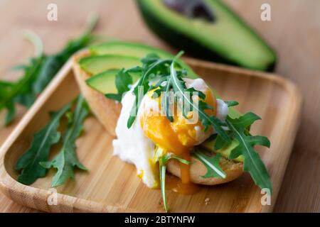 Colazione sana ed elegante con avocado, rucola e toast all'uovo in camicia su una superficie di legno con mezzo avocado e rucola sullo sfondo Foto Stock