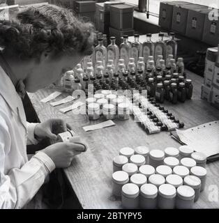 Anni '50, storico, una lavoratrice in un cappotto bianco seduto ad un banco di lavoro etichettare bottiglie di pillole o compresse mediche, presso la casa farmaceutica di produzione e forniture, Arthur H. Cox's di Brighton, Inghilterra, Regno Unito. Il fondatore della società Arthur H. Cox ha inventato il rivestimento delle compresse. Foto Stock