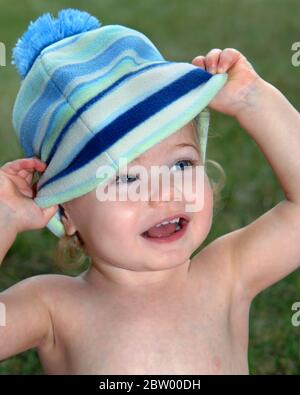 La bambina gioca a peek-a-boo con il suo cappello a righe blu. È sorridente e felice. Foto Stock