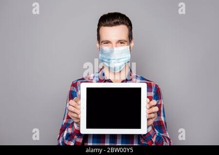 Primo piano ritratto del suo bel ragazzo allegro e attraente che indossa una maschera di protezione camicia controllata che mostra pubblicità schermo tablet isolato su grigio Foto Stock