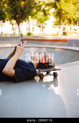 Skater giovane in cuffie arancioni sdraiato sullo skateboard mentre sognando utilizzando il cellulare passare il tempo allo skatepark Foto Stock