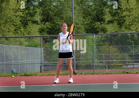 Il giocatore di tennis di Varsity attende di ricevere il servizio durante una partita di tennis. Indossa una gonna da tennis e una maglietta. Il campo è all'aperto. Foto Stock