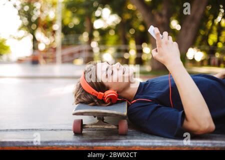 Ritratto di un giovane ragazzo in cuffie arancioni sdraiato sullo skateboard mentre si usa felicemente il cellulare trascorrere del tempo allo skatepark Foto Stock