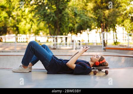 Skater giovane in cuffie arancioni sdraiato su skateboard con cura utilizzando il cellulare con skatepark sullo sfondo Foto Stock