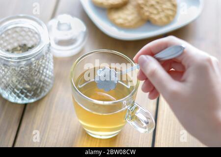 Mano femminile che tiene il cucchiaio a forma di fiore e che agita il tè caldo. Vaso di vetro con tè sciolto e biscotti su tavolo di legno. Foto Stock