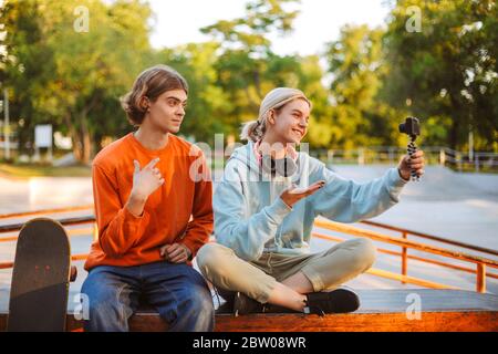 Giovane skater boy in pullover arancione e ragazza con cuffie che registra nuovi video per vlog mentre si passa felicemente tempo insieme allo skatepark Foto Stock