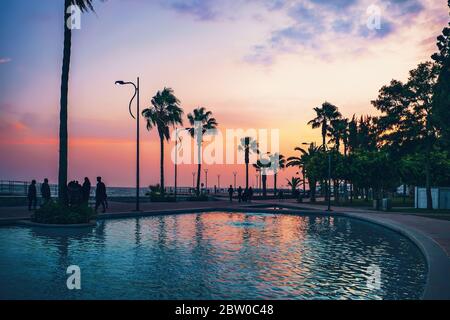 Limassol , Cipro. Sagome di palme e turisti a piedi persone su Molos Park sullo sfondo del cielo tramonto sera. Foto Stock