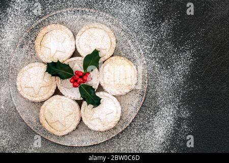 Torte fatte in casa di mince natalizie su un piatto con agrifoglio di bacche invernali e zucchero a velo spolverato su sfondo grigio grunge. Composizione del cibo festivo. Posa piatta, Foto Stock