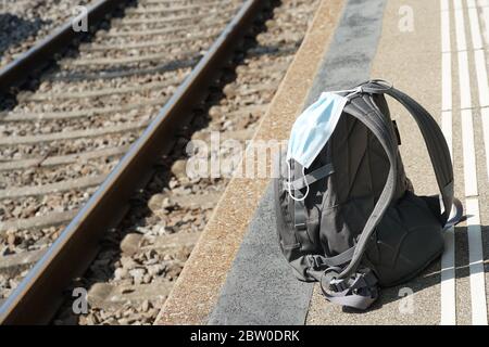 uno zaino con una maschera n95 attaccata su di esso, su una piattaforma della stazione ferroviaria con rotaie sullo sfondo, trasporto al coronavirus covid-19 tempo Foto Stock