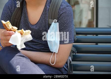 Un adolescente che mangia un breztel, la sua maschera N95 attaccata allo zaino mentre sta mangiando. Regolazione delle abitudini alimentari in coronavirus o COVID-19 Foto Stock