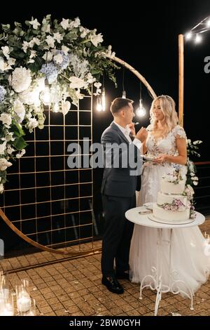 Bella newlywed in piedi vicino all'arco di nozze e assaggiando la loro torta di nozze a tre livelli decorata con fiori Foto Stock
