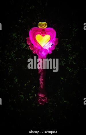 Bacchetta magica a forma di cuore, giocattolo, rosa e gialla, luminosa nell'erba scura di notte. Vista dall'alto, scatto verticale. Foto Stock