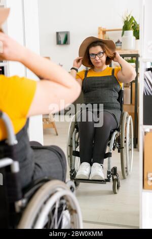Bella donna porta in occhiali seduta in sedia a rotelle davanti allo specchio e indossare un bel cappello prima di passeggiare Foto Stock