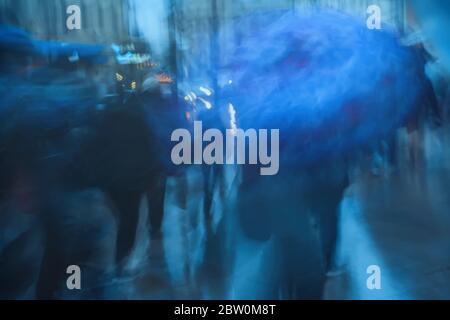 Pioggia fredda sera in città. Sagome di persone sotto gli ombrelloni. Sfondo offuscato astratto in toni blu. Foto Stock