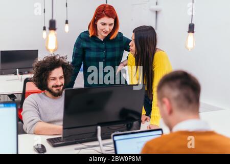 Due giovani donne d'affari accanto a un collega maschile mentre spiega le nuove attività online su un computer in un ambiente di ufficio confortevole. Foto Stock