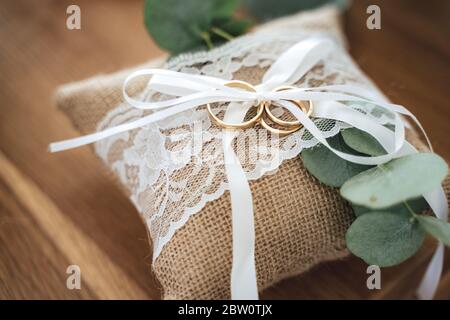 Tre anelli dorati di nozze su piccolo cuscino di burlap decorato con pizzo e nastro bianchi. Sfondo di legno. Concetto di giorno di nozze. Foto Stock