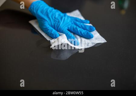 Mano da donna con guanti blu per pulire la superficie del tavolo dell'ufficio domestico con salviette bagnate Foto Stock