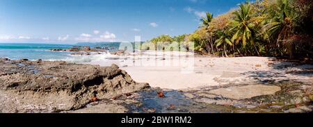 Vista panoramica su una vuota spiaggia di sabbia bianca in Costa Rica con palme e onde blu e chiare che si ondeggia in estate con rocce in avanti Foto Stock