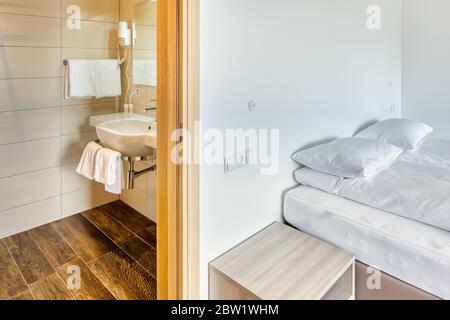 Moderna camera minimalista con bagno chiuso, specchio, cuscino, lavello e asciugamani. Pavimento in piastrelle di legno. Parete bianca senza decorazione. Foto Stock