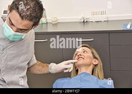 Medico dentale con maschera blu controlla la salute della bocca della sua paziente femminile sdraiata nella poltrona di una moderna clinica dentale. Foto Stock