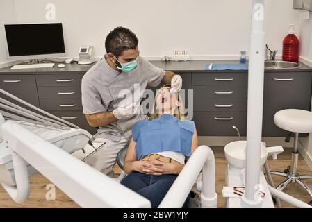 Medico dentale con maschera blu controlla la salute della bocca della sua paziente femminile sdraiata nella poltrona di una moderna clinica dentale. Foto Stock