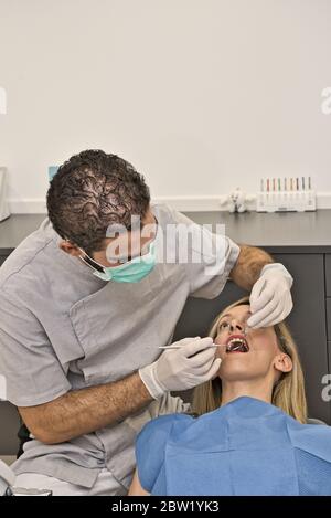 Medico dentale con maschera blu controlla la salute della bocca della sua paziente femminile sdraiata nella poltrona di una moderna clinica dentale. Foto Stock