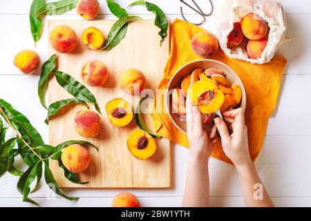 Mani femminili che tagliano pesche dolci fresche. Pesche foglie di frutta intere, mezza pesca, fette di pesca su tavolo da cucina in legno bianco. Ricetta per la preparazione della marmellata di pesche Foto Stock