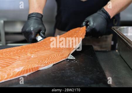 Master shusi con guanti neri igienici pulizia e preparazione di un enorme salmone fresco. Rimozione e peeling della pelle dal pesce. Cucina e cibo conc Foto Stock