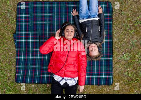 vista dall'alto di due sorelle con occhi chiusi in cuffia, ascolta la musica che si trova su una coperta a scacchi in una foresta Foto Stock