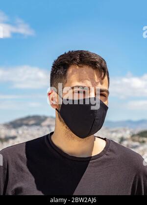 Atene Grecia giorni coronavirus. Giovane uomo che indossa maschera protettiva, paesaggio urbano di Atene e sfondo blu cielo. Foto Stock