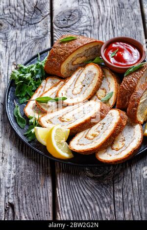 Al forno Formaggi all'Omelet Roll con carne tritata ripieno su un piatto nero con limone, ketchup e prezzemolo su tavola di legno grigio con forchetta dorata e k Foto Stock