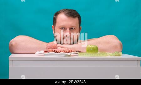 L'attraente giovane uomo grasso in t-shirt casual rende facile la scelta tra mela verde o salsiccia, scegliendo frutta organica sana invece di malsana veloce Foto Stock