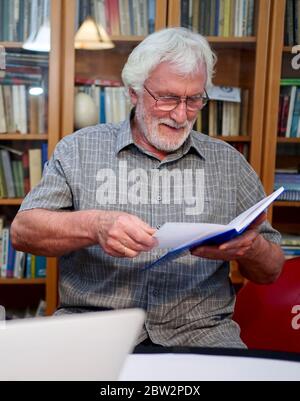 Ritratto di un uomo anziano con bearded bianco con camicia grigia che legge un notebook blu, libro mentre si siede davanti ad un enorme scaffale del libro sullo sfondo. Foto Stock