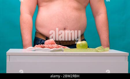 l'uomo con una pancia grassa sceglie tra salsiccia e mela verde. Il concetto di dieta e di volontà. Foto Stock