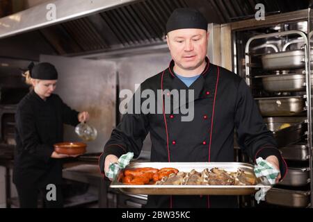 Ritratto di chef sorridente sicuro che lavora in cucina ristorante, ottenendo piatto di carne pronto fuori dal forno Foto Stock