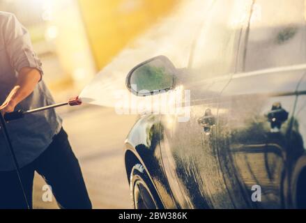 Settore dei trasporti. Uomini caucasici nei suoi 30 anni lavando la sua moderna auto utilizzando una potente macchina idropulitrice. Tema automobilistico. Foto Stock