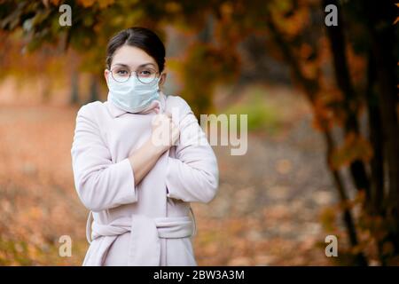 ragazza in una maschera medica viso si avvolge in un cappotto in un giorno di autunno Foto Stock