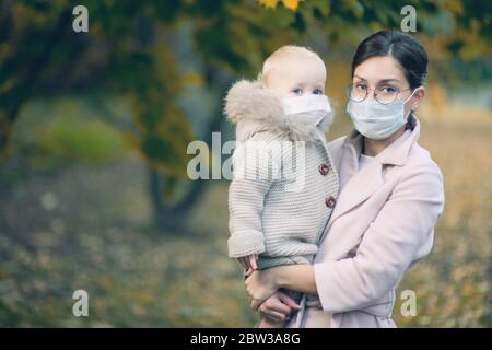 una donna e un bambino piccolo in braccio in maschere mediche di protezione durante l'epidemia. mamma e bambina in maschere mediche nella foresta autunnale. Co Foto Stock