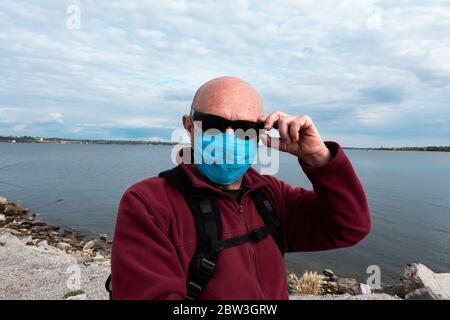 un uomo calvo in un maglione rosso con una maschera medica protettiva e occhiali da sole si trova sulla riva del fiume. Un uomo anziano in un periodo di autoisolamento e q Foto Stock