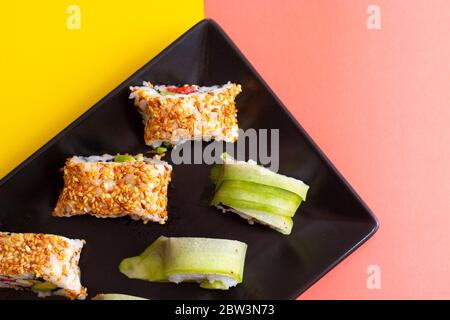 Vista dall'alto di un piatto di sushi con semi di sesamo e un involtino di cetrioli, in una combinazione di vibranti colori rosa e giallo. Foto Stock